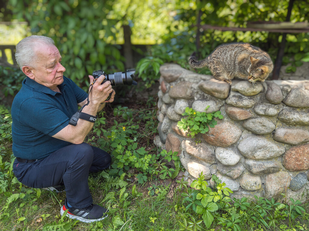 Mężczyzna w niebieskiej koszulce kuca z aparatem Sony, fotografując kota stojącego na kamiennym murku w otoczeniu zieleni. Ujęcie wykonane w słoneczny dzień, w ogrodzie pełnym roślinności. Kot wpatruje się w coś na murze, a fotograf skupia się na uchwyceniu idealnego kadru.
